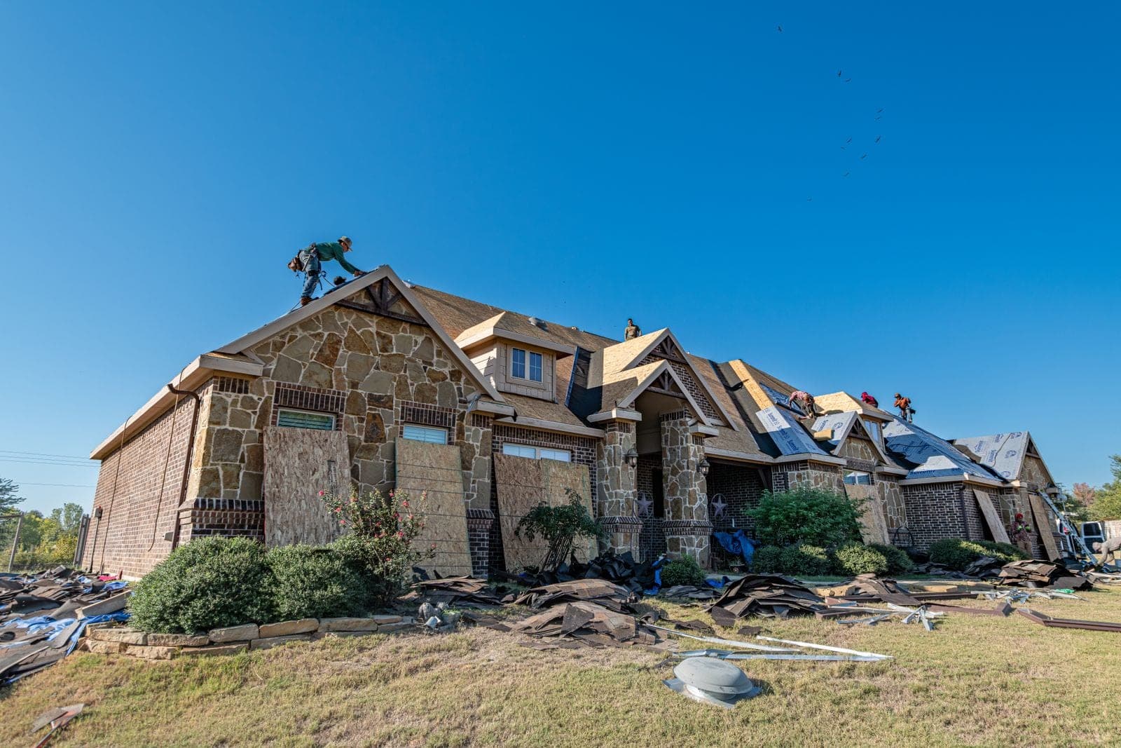 Brick and stone home with roofers and plywood boards and old shingles scattered on the lawn